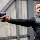 Ein Mann in schwarzer Kleidung und Weste zielt mit einer Pistole in einem industriellen Gebäude.