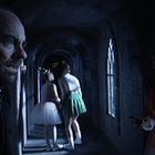 In einem düsteren, schmalen Korridor schaut ein Mann besorgt. Zwei Mädchen in Ballettkleidung umarmen sich, eine Gestalt lurkt rechts.