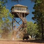Ein hölzerner Aussichtsturm mit einer Treppe führt zu einem kleinen Gebäude. Ein Geländewagen steht unten. Bäume umgeben die Szene.