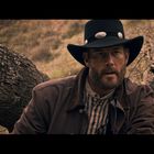 Ein Mann mit einem schwarzen Cowboyhut und brauner Jacke sitzt vor einem gefallenen Baum. Die Landschaft im Hintergrund ist trocken.