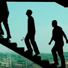 Drei Silhouetten steigen eine Treppe hinauf, vor einem hellblauen Himmel im Hintergrund.