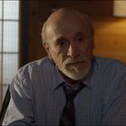 Ein älterer Mann im Hemd sitzt in einem stimmungsvoll beleuchteten Raum mit Holzwänden und einer Lampe im Hintergrund.