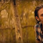 Ein Mann mit bart steht in einem Wald, trägt ein kariertes Hemd, schaut konzentriert. Hintergrund zeigt Bäume.