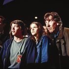 Personen blicken gespannt in dieselbe Richtung. Ein Mann trägt ein Schild mit "Visitor". Szenerie in dunkler Umgebung.