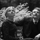 Zwei Personen in historischer Kleidung sitzen auf einer Bank und schauen nach oben. Im Hintergrund sind Pflanzen sichtbar.