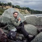 Ein Mann in grüner Jacke sitzt zwischen großen Felsen, hält eine Kamera und blickt überrascht. Im Hintergrund Klippen und Meer.