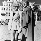 Zwei Personen stehen am Strand vor einem großen Gebäude. Beide tragen Mäntel. Eine Person lächelt.