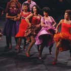 Tanzende Menschen in farbenfrohen Kleidern, energische Bewegungen auf einer Bühne.