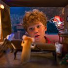 Ein Junge mit lockigem Haar schaut neugierig in ein Puppenhaus mit einer aufrechtstehenden Maus vor einem Sofa.