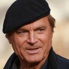 Ein Mann mit schwarzer Mütze und dunkler Jacke, lächelnd, vor verschwommenem Hintergrund.