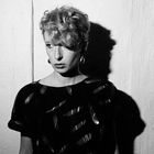 Keine erkennbaren Texte. Schwarz-Weiß-Foto einer Person mit lockigem Haar und ernstem Ausdruck, vor einer Wand.