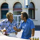 Zwei Männer in gemusterten Hemden unterhalten sich; im Vordergrund Dosen Olympian-Bier auf dem Autodach.