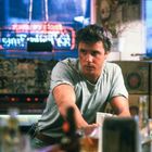 Leuchtendes Schild mit Neontext im Hintergrund, Mann in grauem T-Shirt sitzt an einer Bar, diverse Flaschen auf dem Tresen.