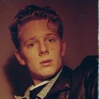 Es gibt keinen Text. Ein junger Mann schaut ernst, trägt eine Lederjacke. Rechts ist ein abstraktes, buntes Muster.