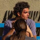 Zwei Personen sitzen draußen. Der Mann mit Locken hat blaue Flecken im Gesicht und sieht nach rechts. Kein Text sichtbar.