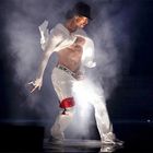 Ein Tänzer in weißer Kleidung, umgeben von Rauch, unter dramatischem Licht. Er trägt einen schwarzen Hut.