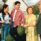 Ich kann den Text nicht erkennen. Vier Personen stehen in einem Garten. Zwei haben Rucksäcke, eine ältere Frau schaut überrascht.