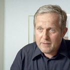 Ein Mann mit grauem Haar und Bartstoppeln trägt ein dunkles Hemd. Hinter ihm ein gerahmtes Bild an der weißen Wand.