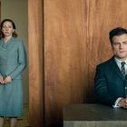 Keine Texte erkennbar. Eine Frau in grauem Kostüm steht, ein Mann in Anzug sitzt an einem Holztisch. Beides vor Holzpaneelen.