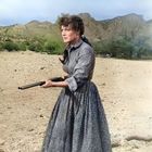 Eine Frau in einem langen Kleid hält ein Gewehr in einer Wüstenlandschaft mit Bergen im Hintergrund.