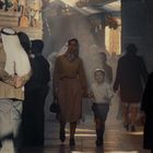 Mehrere Menschen in traditioneller Kleidung gehen durch einen belebten Markt, umgeben von bunten Ständen.