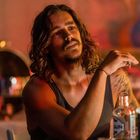 Ich kann die Texte im Bild nicht erkennen. Ein Mann mit langen Haaren sitzt entspannt an einem Tisch mit Flaschen.