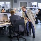 Ein Büro mit mehreren Personen, die an Schreibtischen arbeiten. Zwei Männer im Vordergrund, einer sitzt, der andere lehnt sich.