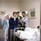 Vier Männer stehen um ein Bett, darauf eine Person mit eingegipstem Fuß. An der Wand hängen zwei Landschaftsgemälde.