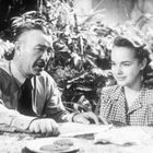 Ein Mann und eine junge Frau sitzen an einem Tisch im Freien, umgeben von Pflanzen, und scheinen vertieft in ein Gespräch zu sein.