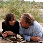 Keine sichtbaren Texte. Eine Frau und ein Mann liegen am Ufer auf einer Decke, umgeben von Pflanzen.