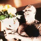 Es gibt keinen Text. Eine gruselige Figur mit verzerrtem Gesicht hält einen Strauß gelber Blumen in den Händen.