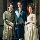 Drei Personen in historischen, eleganten Gewändern stehen nebeneinander in einem opulenten Raum.