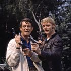 Zwei Männer im Freien, beide halten Pistolen; einer trägt eine helle Jacke, der andere eine schwarze Lederjacke.