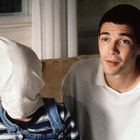 Ein Mann in weißem Pullover sitzt neben einer Person mit Stoff über dem Kopf, die gestreifte Latzhose trägt.