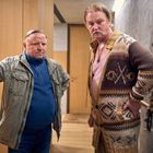 Zwei Männer stehen in einem Raum mit Holztüren. Der linke trägt eine graue Jacke, der rechte einen gemusterten Cardigan.