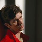 Ich kann keine Texte im Bild erkennen. Ein Mann mit braunem Haar schaut nachdenklich zur Seite. Er trägt eine rote Jacke.