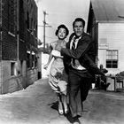 Text auf einem Schild: "RECEIVING". Eine Frau und ein Mann rennen durch eine Gasse, er hält ihre Hand.