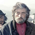 Ein Mann mit zerzaustem Haar und Bart schaut ernst. Zwei weitere Männer sind im Hintergrund, einer mit Pfeife, der andere unscharf.