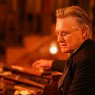 Ein älterer Mann mit Brille sitzt in warm beleuchtetem Raum, schaut nach rechts, eine brennende Lampe im Vordergrund.