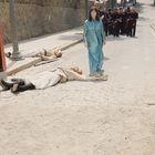 Eine Frau in einem blauen Kleid geht auf einer Straße, vorbei an zwei liegenden Personen und einer Gruppe in schwarzer Kleidung.