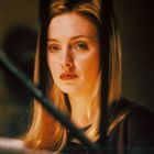 Keine Texte zu sehen. Eine Frau mit blonden Haaren schaut ernst zwischen dunklen Streben hindurch.