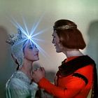 Person in Silberkrone mit Lichtstrahl, hält Hände einer Person in Rot mit Stirnband, vor diffuser Kulisse.