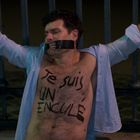 "Te suis un enculé" steht auf dem Oberkörper eines Mannes, der mit Klebeband gefesselt ist. Seine Arme sind ausgestreckt.