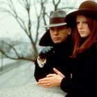 Zwei Personen stehen draußen an einer winterlichen Mauer. Sie tragen Hüte und Mäntel und schauen ernst in die Kamera.