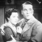 Ein Schwarzweißfoto zeigt ein Paar in inniger Umarmung, ernst in die Ferne blickend.