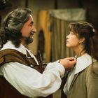 Ein Mann, der historische Kleidung trägt, richtet einer jungen Frau den Kragen. Beide schauen sich an.