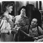 Drei Personen in traditioneller Kleidung, eine Frau hält ein Tuch, ein Mann sitzt mit misstrauischem Blick, Küche im Hintergrund.