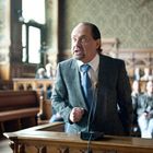 Ein Mann in Anzug vor einem Mikrofon in einem Gerichtssaal; Publikum sitzt im Hintergrund.