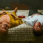Zwei Personen liegen auf einem Bett: links in einem goldenen Gewand, rechts in einem weißen Hemd.
