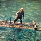 Ein Mann steht auf einem einfachen Floß aus Baumstämmen und treibt auf einem Fluss, umgeben von grünem Wasser und Pflanzen.